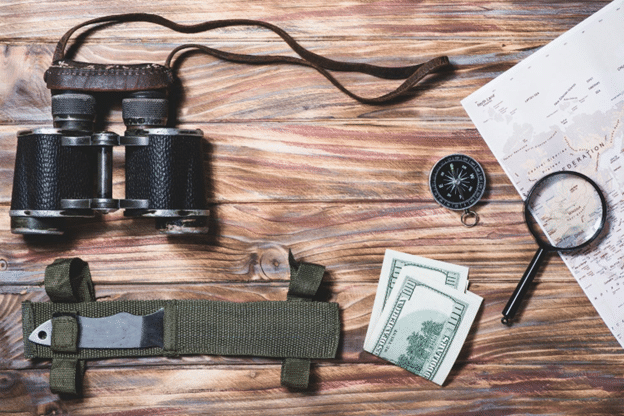 Travel essentials including binoculars, compass, map, magnifying glass, utility knife, and cash on a wooden surface.